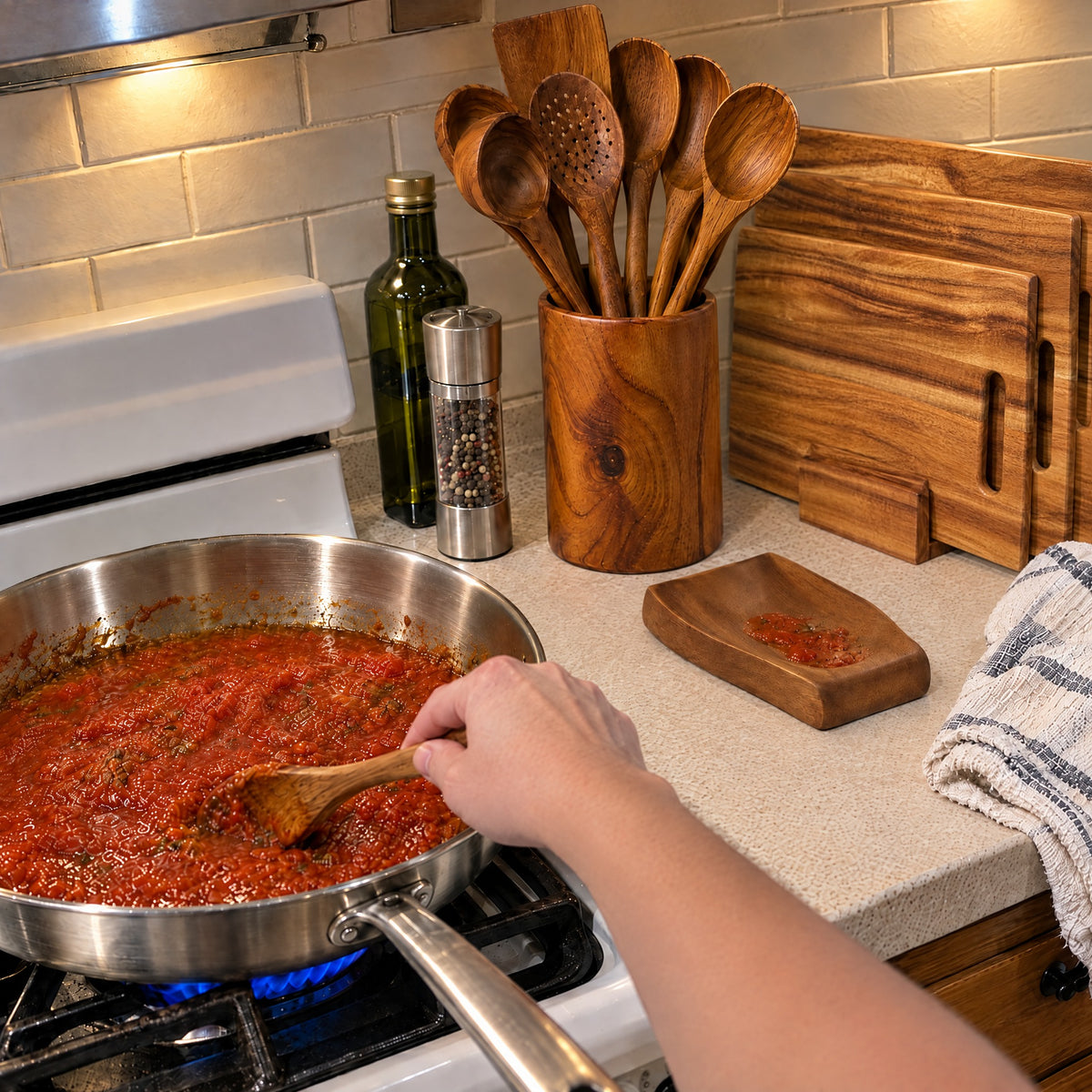 Cooking with a teak spoon in a stainless steel pan, with the utensil holder, spoon rest and three acacia cutting boards on the counter