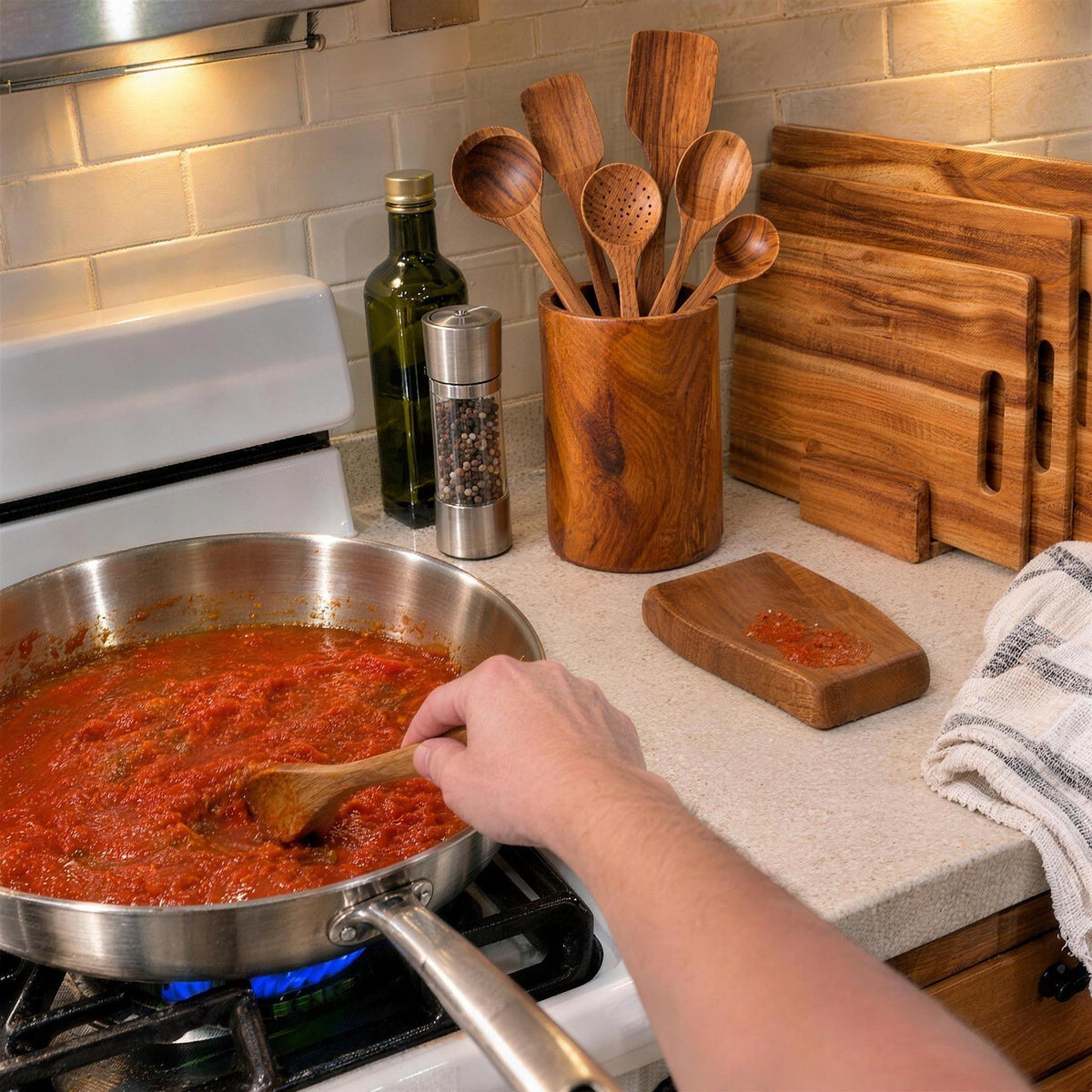 Cooking with an acacia spoon in a stainless steel pan, with the utensil holder, spoon rest and three acacia cutting boards on the counter